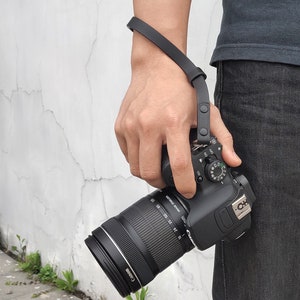 Pu&ograve; includere: Una fotocamera nera con un obiettivo nero attaccato a un cinturino da polso nero. La fotocamera ha un'impugnatura nera e un pulsante nero nella parte superiore.