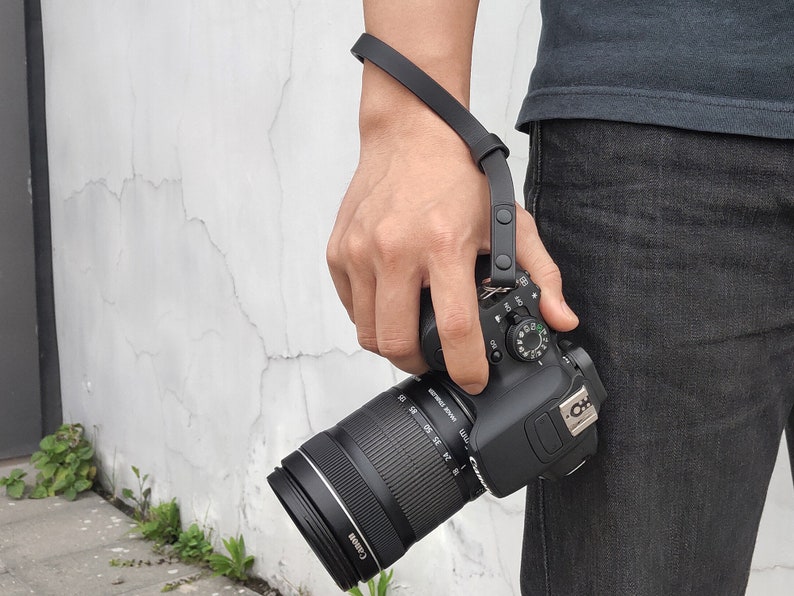 Pu&ograve; includere: Una fotocamera nera con un obiettivo nero attaccato a un cinturino da polso nero. La fotocamera ha un'impugnatura nera e un pulsante nero nella parte superiore.