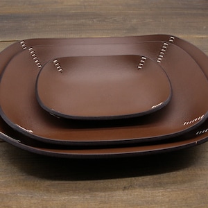 May include: Three brown leather trays of different sizes, nested together. The trays have a rounded rectangular shape and are stitched with white thread around the edges.