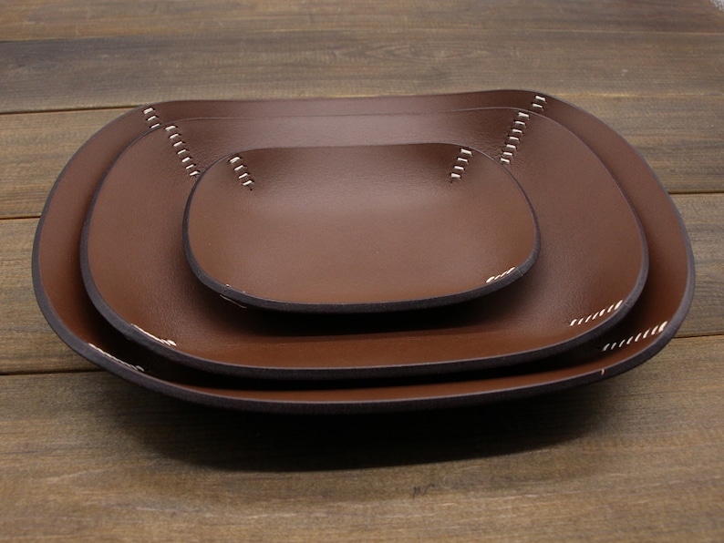 May include: Three brown leather trays of different sizes, nested together. The trays have a rounded rectangular shape and are stitched with white thread around the edges.