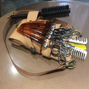 Puede incluir: Un cintur&oacute;n de herramientas de peluquer&iacute;a de cuero color canela con una correa para el hombro, que contiene tijeras, peines y pinzas para el pelo. Un cepillo redondo con mango negro descansa encima. Las herramientas est&aacute;n dispuestas sobre una superficie marr&oacute;n.