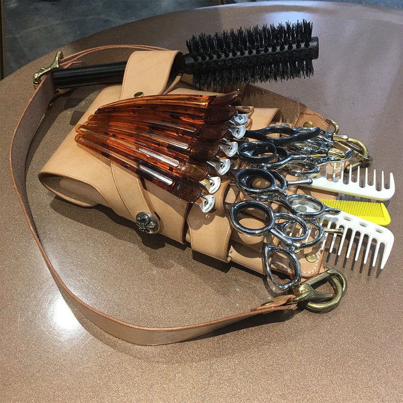 Puede incluir: Un cintur&oacute;n de herramientas de peluquer&iacute;a de cuero color canela con una correa para el hombro, que contiene tijeras, peines y pinzas para el pelo. Un cepillo redondo con mango negro descansa encima. Las herramientas est&aacute;n dispuestas sobre una superficie marr&oacute;n.