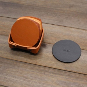 Puede incluir: Un juego de seis posavasos de cuero marr&oacute;n en un soporte de cuero. Los posavasos est&aacute;n apilados uno encima del otro y el soporte est&aacute; abierto. Un solo posavasos de cuero negro con la palabra "BLW" en relieve est&aacute; sobre una superficie de madera.