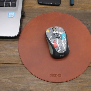 May include: A round brown leather mouse mat with a black mouse featuring a cartoon design on top. The mouse mat has the word "SHENG" embossed on it.