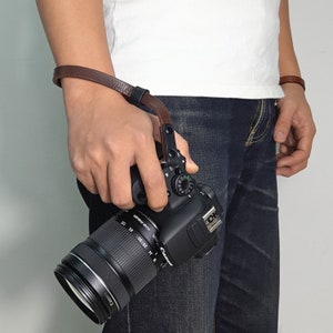 May include: A brown leather camera wrist strap with a metal buckle attached to a black DSLR camera with a large lens.