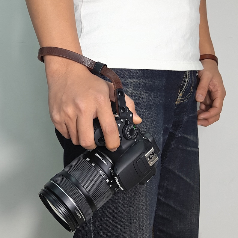 May include: A brown leather camera wrist strap with a metal buckle attached to a black DSLR camera with a large lens.