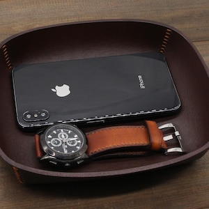 May include: A brown leather valet tray with a black iPhone and a black wristwatch with a brown leather band. The watch has a silver buckle and a white face with black hands and numbers.