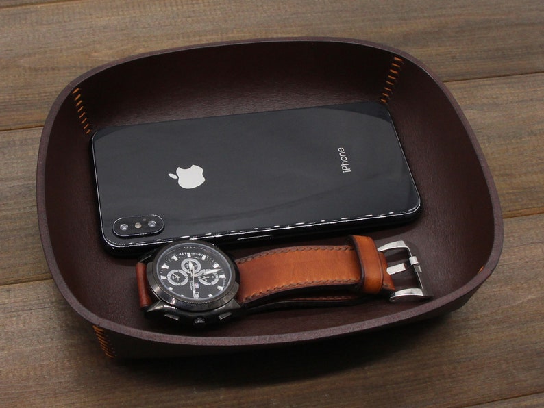May include: A brown leather valet tray with a black iPhone and a black wristwatch with a brown leather band. The watch has a silver buckle and a white face with black hands and numbers.