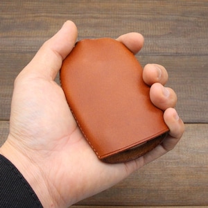 May include: A brown leather key holder with a curved shape. The key holder is held in a hand, showcasing its smooth texture and design. The stitching is visible along the edges, highlighting the craftsmanship.