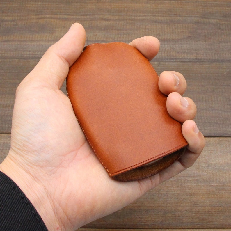 May include: A brown leather key holder with a curved shape. The key holder is held in a hand, showcasing its smooth texture and design. The stitching is visible along the edges, highlighting the craftsmanship.