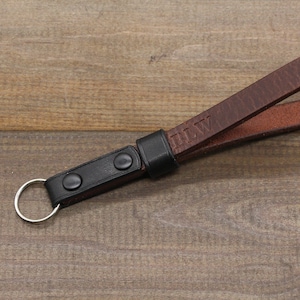 May include: A brown leather camera strap with a black leather adjustable strap and a metal ring. The strap is personalized with the initials "BLW".