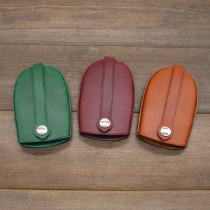 May include: Three leather key holders in green, burgundy, and tan. Each has a loop and a silver-coloured snap closure. The key cases are arranged on a wooden surface, showcasing their colours and design.