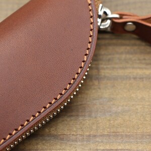 May include: A brown leather zippered pouch with a stitched edge and a leather strap with a metal buckle.