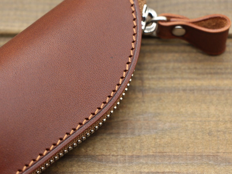 May include: A brown leather zippered pouch with a stitched edge and a leather strap with a metal buckle.