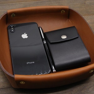 May include: A brown leather tray with a black leather wallet, a black smartphone with the word "iPhone" on the screen, and a silver pen.