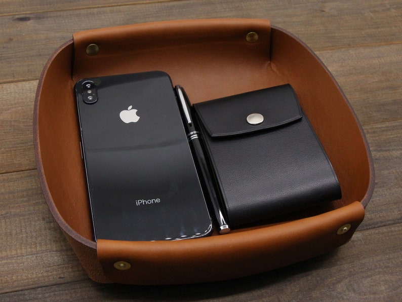 May include: A brown leather tray with a black leather wallet, a black smartphone with the word "iPhone" on the screen, and a silver pen.