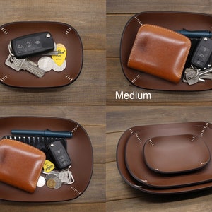 May include: Three leather trays in different sizes: small, medium, and large. The trays are brown and have a stitched edge. The small tray has a key and a few coins in it. The medium tray has a brown leather wallet and keys. The large tray has a brown leather wallet, keys, and a pen.
