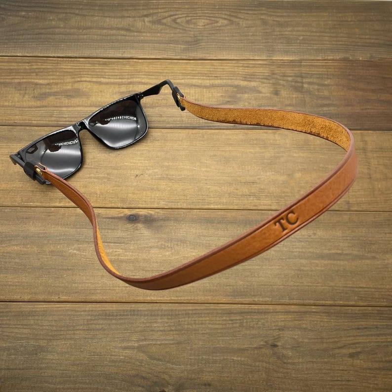 May include: Black sunglasses with a brown leather strap. The strap is engraved with the initials "TC". The sunglasses are resting on a wooden surface.