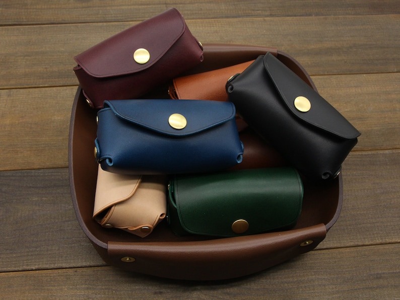 May include: A collection of six leather coin purses in various colors, including burgundy, brown, blue, green, and beige. Each purse has a brass button closure and is displayed in a brown leather tray.