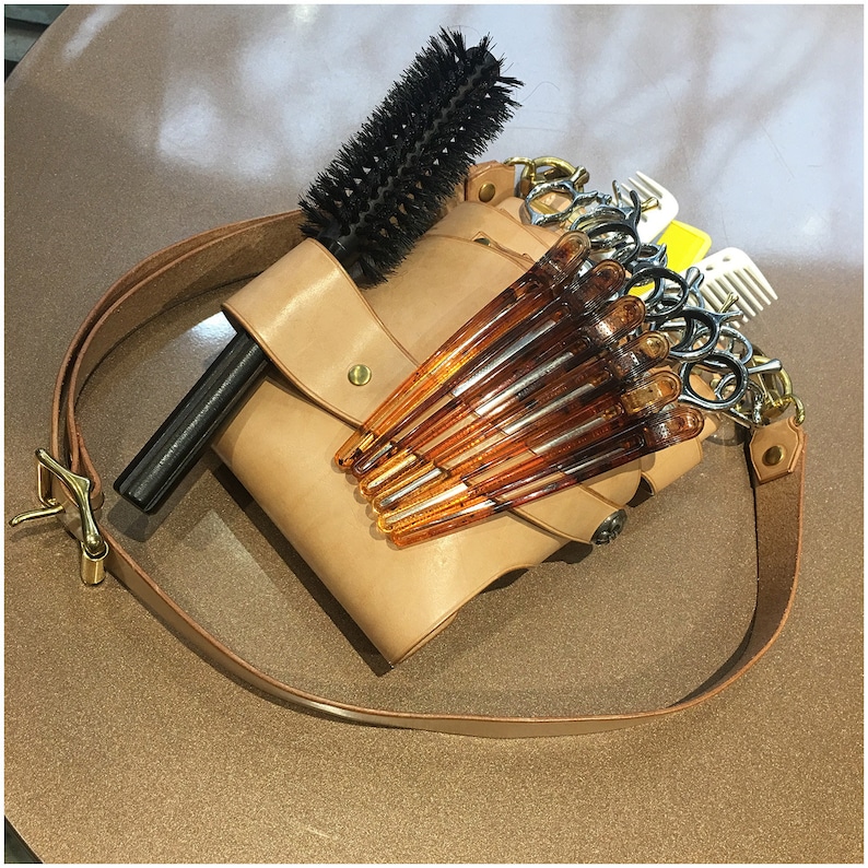 Puede incluir: Un cintur&oacute;n de herramientas de peluquer&iacute;a de cuero color canela con un cepillo redondo de cerdas negras, pinzas para el pelo de color &aacute;mbar y varios pares de tijeras. El cintur&oacute;n tiene una correa de cuero marr&oacute;n con una hebilla dorada.