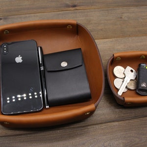 May include: Two brown leather trays with a black phone, pen, wallet, keys, and coins. The larger tray has a black phone, pen, and wallet. The smaller tray has keys and coins.