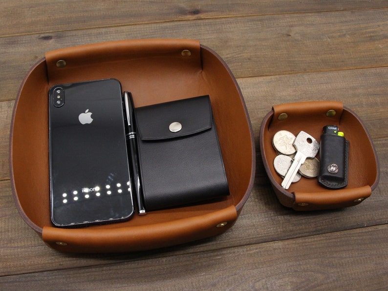 May include: Two brown leather trays with a black phone, pen, wallet, keys, and coins. The larger tray has a black phone, pen, and wallet. The smaller tray has keys and coins.