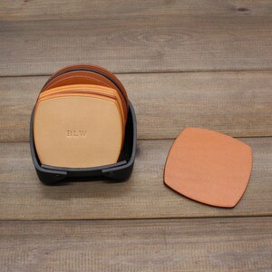 Puede incluir: Un juego de seis posavasos de cuero marr&oacute;n con un soporte negro. Los posavasos est&aacute;n apilados y el posavasos superior tiene las iniciales "BLW" grabadas.