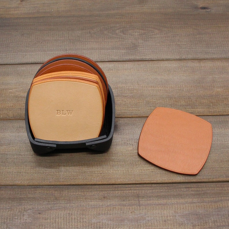 Puede incluir: Un juego de seis posavasos de cuero marr&oacute;n con un soporte negro. Los posavasos est&aacute;n apilados y el posavasos superior tiene las iniciales "BLW" grabadas.