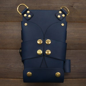 Puede incluir: Una funda de cuero azul marino con herrajes met&aacute;licos dorados. La funda presenta m&uacute;ltiples correas, remaches decorativos y dos anillas en D en la parte superior. El dise&ntilde;o es sim&eacute;trico y parece hecho a mano.