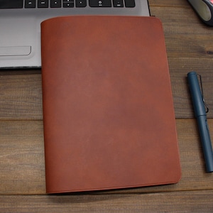 May include: A brown leather notebook cover with a smooth, textured surface. The cover is closed and lying flat on a wooden surface.