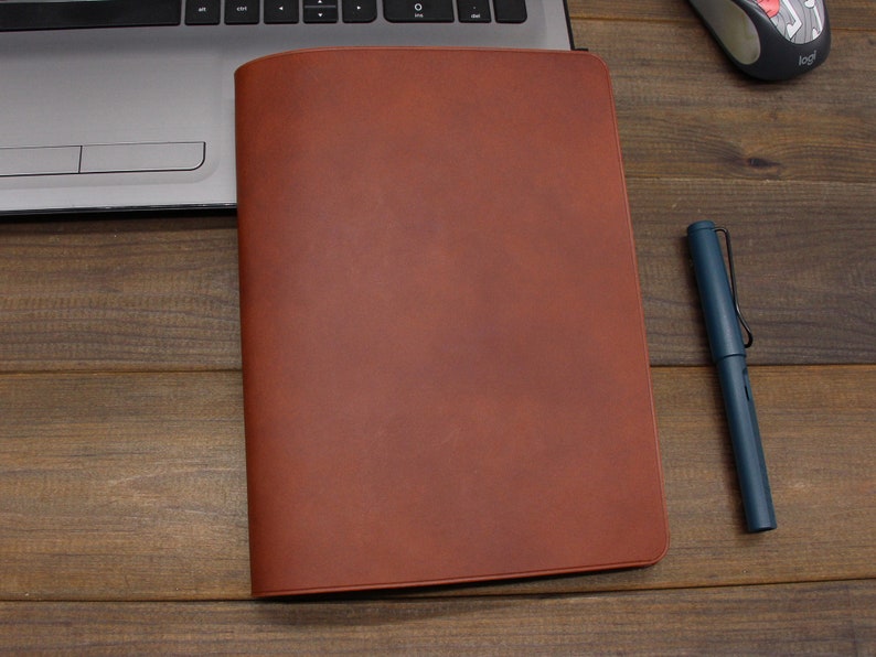 May include: A brown leather notebook cover with a smooth, textured surface. The cover is closed and lying flat on a wooden surface.