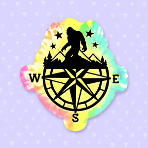 May include: A yellow, green, and pink tie-dye sticker with a black compass and a silhouette of Bigfoot standing on a mountain range. The compass points are labeled W, E, and S.
