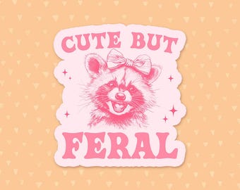 Op de afbeelding: Roze sticker met een schattige cartoonwasbeer met een roze strik en de tekst "Cute But Feral".