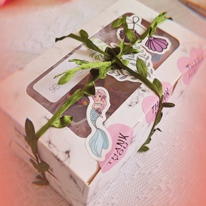 May include: A white gift box with a marble pattern, decorated with green leaves, a pink heart sticker that says "Thank You", and a mermaid sticker. The box is tied with a green ribbon.