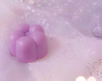 May include: A small, solid, light purple flower-shaped object with rounded petals. The item has a smooth, matte texture and a uniform pastel colour. It is positioned on a white, circular, lacy paper doily with intricate cut-out patterns.