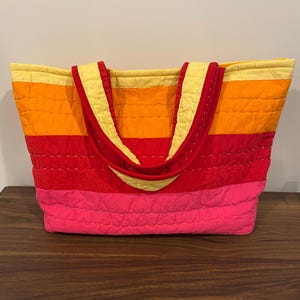 May include: A quilted tote bag with horizontal stripes in shades of yellow, orange, red, and pink. The bag has red handles and a yellow interior lining. The bag is made of a soft, quilted material.
