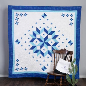 Könnte beinhalten: Eine weiße Steppdecke mit einem blauen und hellblauen Sternenmuster. Die Steppdecke hängt an einer Wand mit einem Schaukelstuhl und einer Kakteenpflanze davor. Der Stuhl hat eine schwarze Tragetasche mit dem Text "Sarah's Softies" darauf.