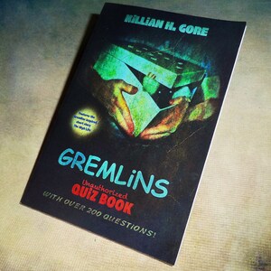 May include: A black and white book cover with the title "Gremlins Unauthorized Quiz Book" and the text "With over 200 questions!" The cover features a hand holding a box with a green and white design.