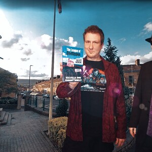 May include: Two people are standing outside holding copies of the book "The League of Gentlemen" by  KILLING IS HARD,  PROVEN BY MURDER. The book cover is blue with a white house and the title in black text.