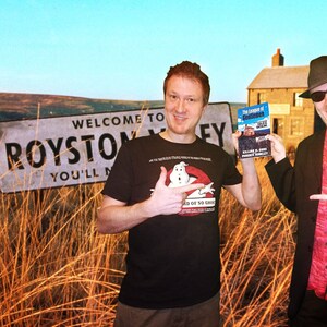 May include: Two people stand in a field of tall grass. One person is wearing a black t-shirt with a Ghostbusters logo and is holding a book titled "The Legend of  Godmanchester". The other person is wearing a black suit, a red shirt, and a black hat with sunglasses. They are both pointing to the sign that reads "Welcome to Royston Valley You'll Never Leave".