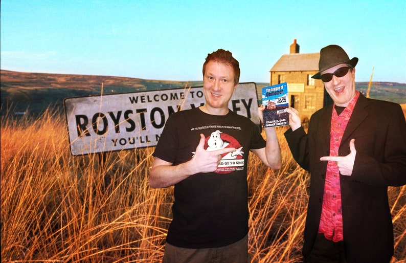 May include: Two people stand in a field of tall grass. One person is wearing a black t-shirt with a Ghostbusters logo and is holding a book titled "The Legend of  Godmanchester". The other person is wearing a black suit, a red shirt, and a black hat with sunglasses. They are both pointing to the sign that reads "Welcome to Royston Valley You'll Never Leave".