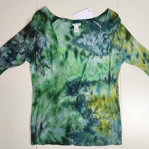 May include: A green, blue, and yellow tie-dye cardigan with a V-neck, short sleeves, and button closure. The sweater has two front pockets and a ribbed texture. A white tag is attached near the neckline.
