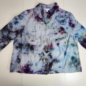 May include: A light-colored denim jacket with a tie-dye pattern in shades of blue, purple, and green. The jacket has a button-up front, two chest pockets, and long sleeves. The jacket is laid flat on a white surface.