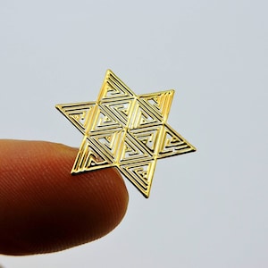 Pode incluir: Uma estrela dourada de seis pontas com um design geométrico de triângulos interligados. A estrela é segurada entre dois dedos contra um fundo branco. O padrão intrincado da estrela cria um efeito delicado e vazado.