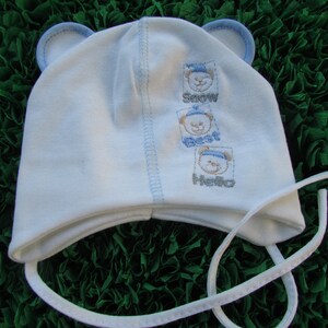 May include: White baby hat with blue trim and bear embroidery. The embroidery says "Snow", "Best", and "Hello".