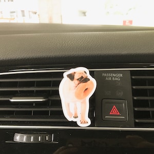 May include: A car air freshener featuring a pug dog with a donut in its mouth. The air freshener is attached to the air vent in a car. The text "PASSENGER AIR BAG" is visible on the dashboard.