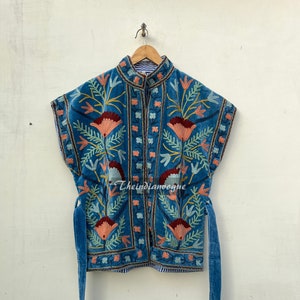 May include: A blue velvet waistcoat with floral embroidery. The waistcoat has a mandarin collar and a button fastening. The waistcoat has two long ties that hang down the sides.