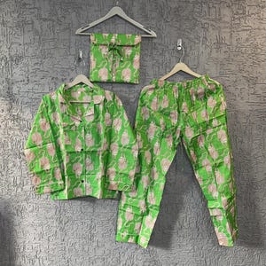 Puede incluir: Un conjunto de pijama verde con un patrón rosa repetido, que incluye una camisa con botones, pantalones y una bolsa con cordón a juego. El conjunto se muestra en perchas contra una pared gris texturizada.
