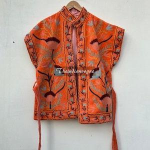 May include: A bright orange sleeveless jacket with intricate floral embroidery in shades of blue, green, and red. The jacket has a mandarin collar and ties at the waist.