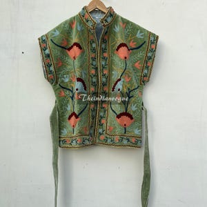 Könnte beinhalten: Eine grüne, bestickte Weste mit floralem Design. Die Weste hat einen hohen Kragen und kurze Ärmel. Die Blumenstickerei weist orange, rote und blaue Akzente auf. Die Weste hängt an einem Holzkleiderbügel vor einer weißen Wand. Theindianvogue.